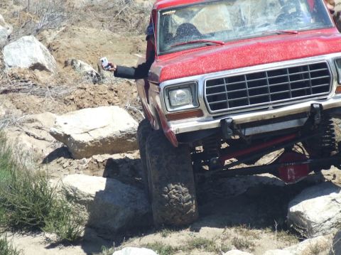 1978 Ford Bronco - A red truck is shown off-roading over large rocks, with the driver holding a phone out the window.