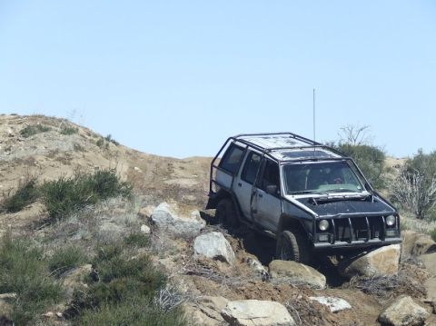 1978 Ford Bronco - An off-road SUV with a protective roll cage navigates a challenging rocky trail under a clear blue sky.