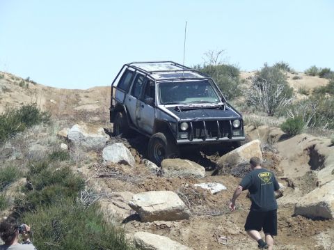 1978 Ford Bronco - A modified Jeep Cherokee XJ is shown tackling a challenging off-road course with large rocks and steep inclines.