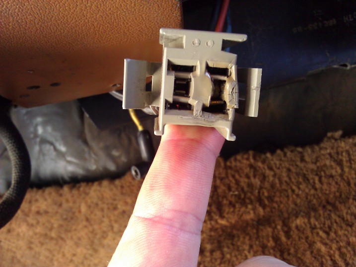 Close-up of a gray electrical connector being held by a finger, with vehicle interior parts in the background