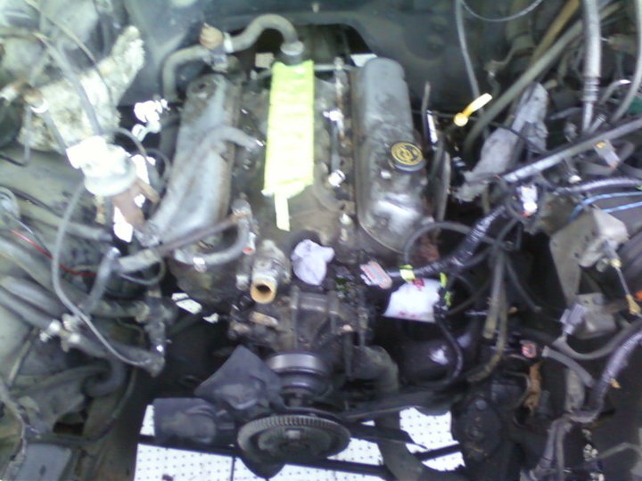 Engine bay showing a V8 engine with various hoses and wiring. A yellow marker is on the engine