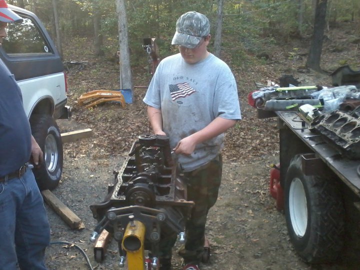 Guy working on engine block next to a white Ford Bronco