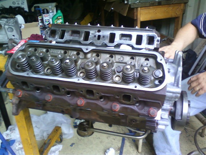 Ford V8 engine block with cylinder heads removed, showing valve springs and pushrods. Engine is on a yellow stand