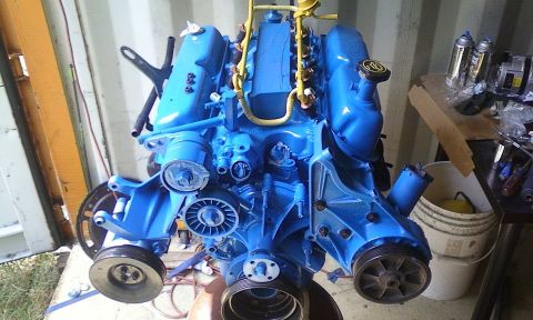 1989 Ford Bronco - A blue engine assembly is shown, with various pulleys and hoses attached, against a backdrop of a shipping container.