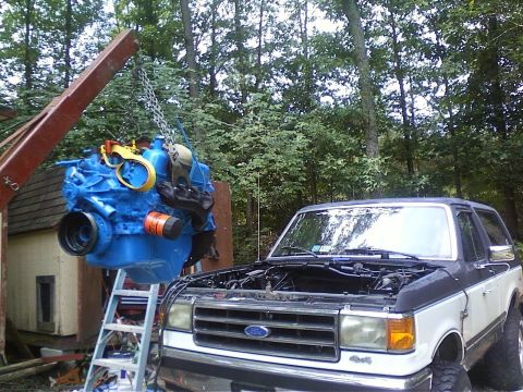 1989 Ford Bronco - A blue engine is being lowered into a two-tone Ford Bronco with its hood open.