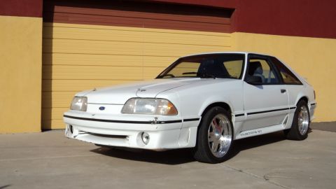 1989 Ford Mustang - This white Ford Mustang GT is parked outside a building with a yellow and red garage door.