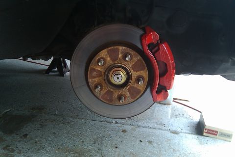 1996 Ford Probe - Close-up of a car's brake assembly, showing a rusty rotor and a red caliper, with the vehicle elevated on jack stands.
