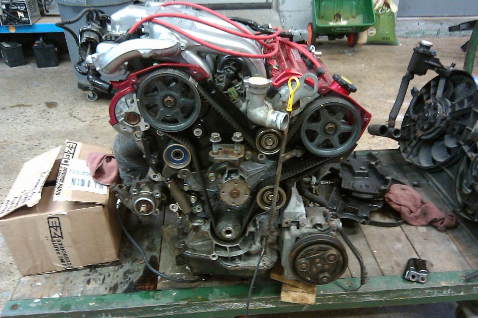 Engine on a stand, showing timing belt and pulleys. Box of parts nearby