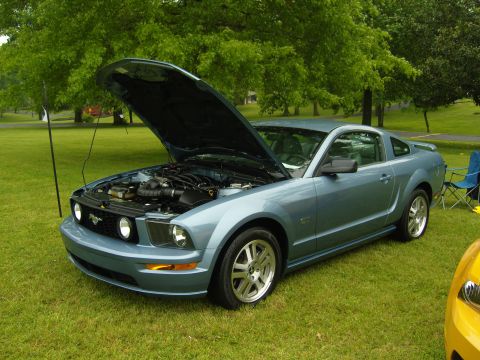 2007 Ford Mustang - A blue 2007 Ford Mustang coupe is displayed with its hood open, parked on a grassy lawn with trees in the background.