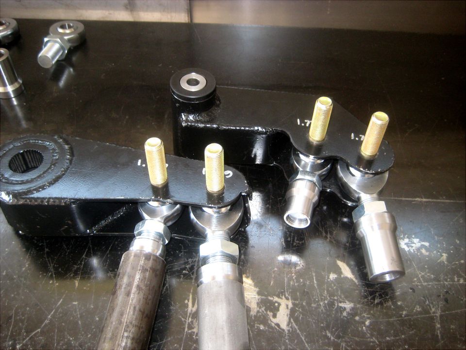 Black metal steering linkage parts with brass pins and silver ball joints on a dark surface