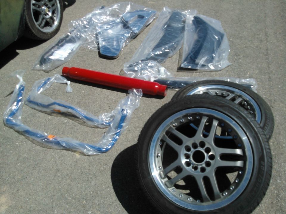 Bagged trim parts, a red bar, and an aftermarket wheel with tire on pavement