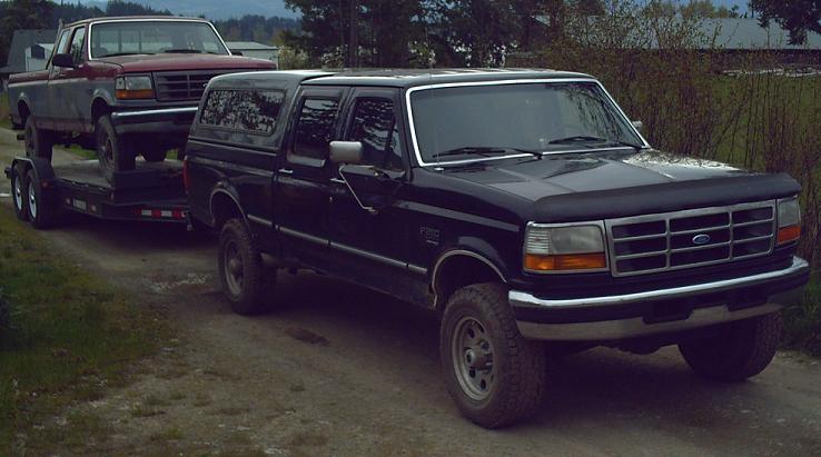 Black Ford crew cab pickup with camper shell towing a red Ford truck on a flatbed trailer