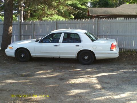 1998 Ford Crown Victoria - A white 1998 Ford Crown Victoria sedan is parked outdoors next to a wooden fence.