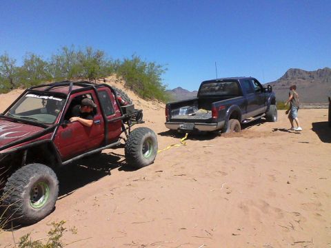 1981 Ford F-150 - A red off-road buggy tows a blue pickup truck that is stuck in the sand dunes under a clear blue sky.