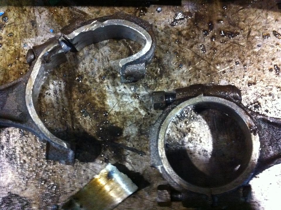 Two broken engine connecting rods on a dirty workbench. One rod is in two pieces, the other is cracked