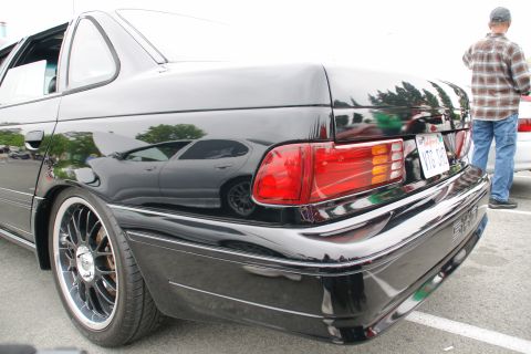 1993 Ford Taurus - A black sedan with aftermarket wheels is viewed from the rear, reflecting trees and another car.