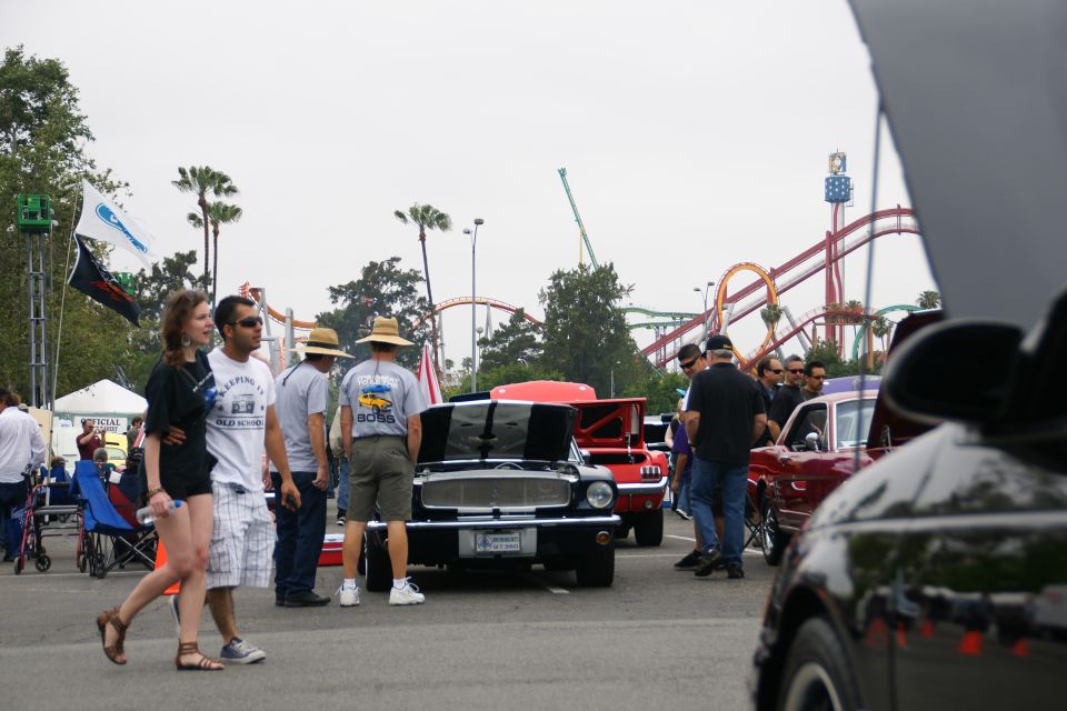People milling around classic cars at an outdoor event with roller coasters in the background