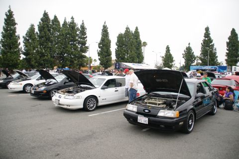 1993 Ford Taurus - A line of cars, including a black 1993 Ford Taurus with its hood open, are parked at an outdoor event.