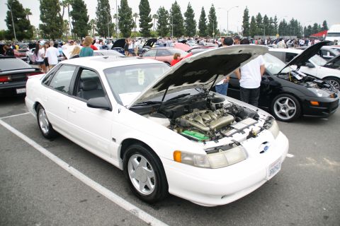 1993 Ford Taurus - A white sedan is parked at an outdoor event with its hood open, revealing the engine compartment.