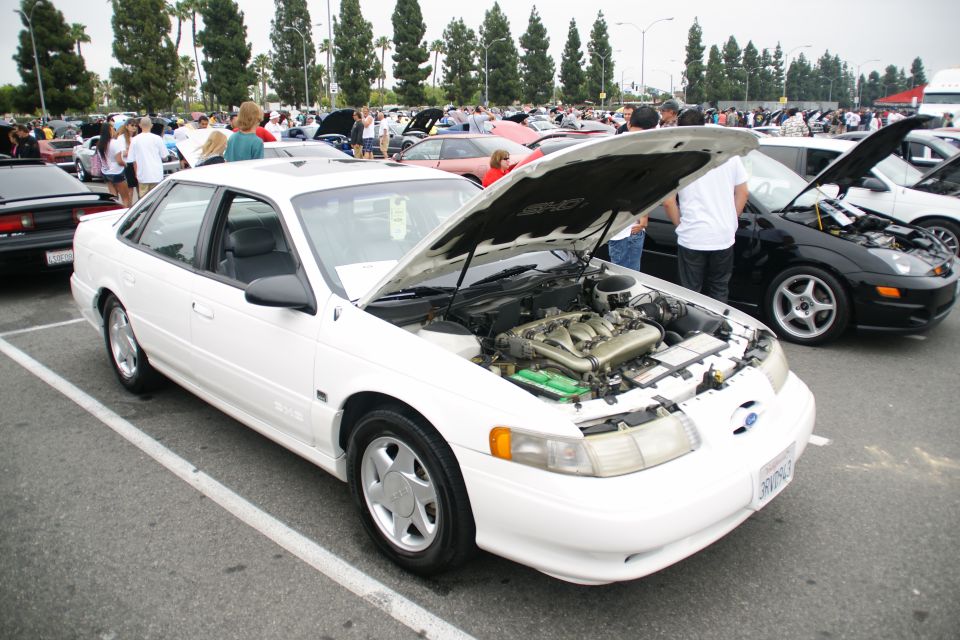 White sedan with hood open, showing engine. Parked at an outdoor car show