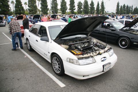 1993 Ford Taurus - A white sedan with its hood open at an outdoor car show, parked next to a black car.