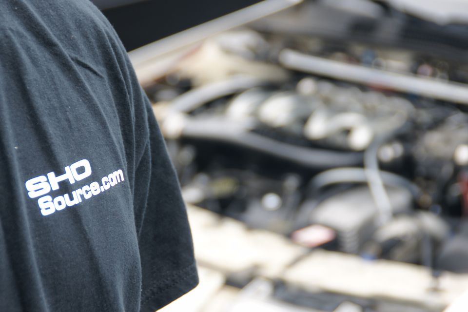 Person wearing black t-shirt with "SHO Source.com" logo, engine bay in background