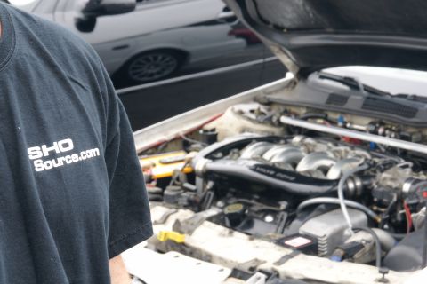 1993 Ford Taurus - The engine bay of a car is shown, with a t-shirt displaying "SHO Source.com" in the foreground. Another vehicle is partially visible in the…