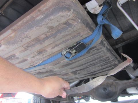 1964 Ford Ranchero - Vehicle fuel tank secured with a ratchet strap, showing underside components and a hand.