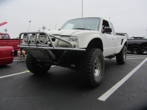 1964 Ford Ranchero - A modified white pickup truck sits in a parking lot, with a red truck visible to the left.