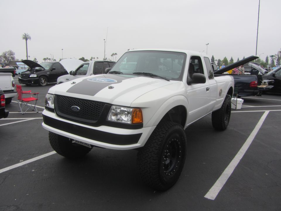 White Ford Ranger pickup with black racing stripes on hood, lifted suspension, and off-road tires. Other vehicles are parked nearby