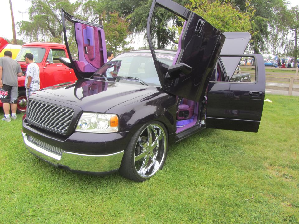 Dark purple Ford F-150 with lambo doors open, purple interior, and chrome rims. Red pickup truck in background