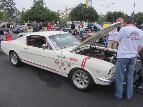1964 Ford Ranchero - A white 1965 Ford Mustang is displayed with its hood open, surrounded by onlookers at an outdoor event.