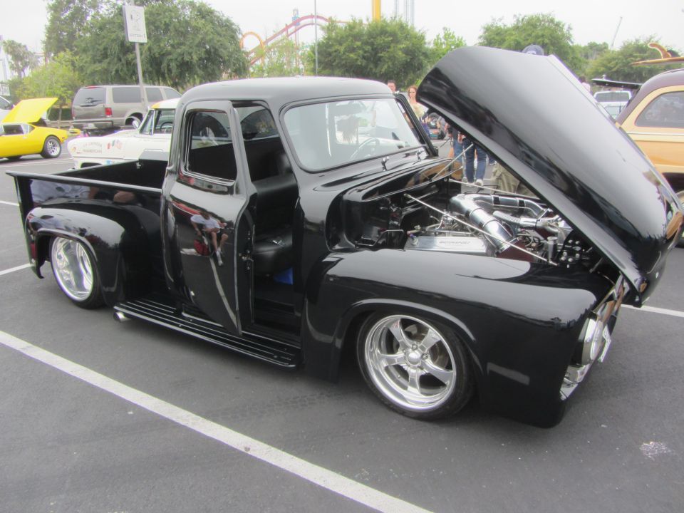 Black 1955 Ford F-100 pickup truck with custom wheels and open hood showing engine