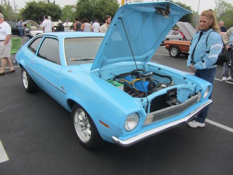1964 Ford Ranchero - A woman stands by a light blue coupe with its hood open, revealing the engine. Several other vehicles and people are visible in the backgro…