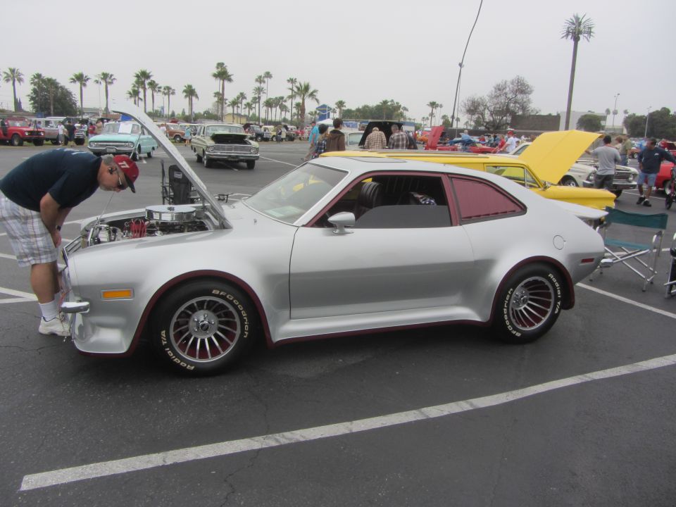 Silver 1974 Ford Pinto coupe with red trim, hood open, guy looking at engine. Other cars in background