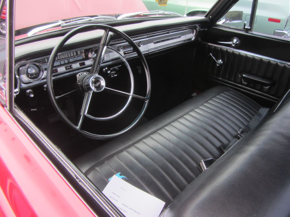 Red 1964 Ford Ranchero interior with black vinyl bench seat, dashboard, and steering wheel. A document is on the seat