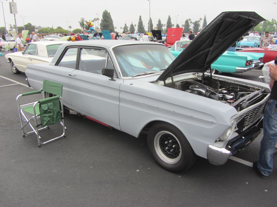 Light gray 1964 Ford Ranchero with hood open, black racing wheels, and a green folding chair beside it
