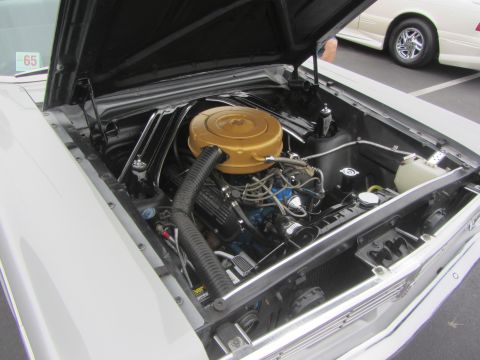1964 Ford Ranchero - The engine bay of a classic vehicle, a gold air cleaner and a visible '65 sticker on the windshield. Another vehicle is parked nearby.