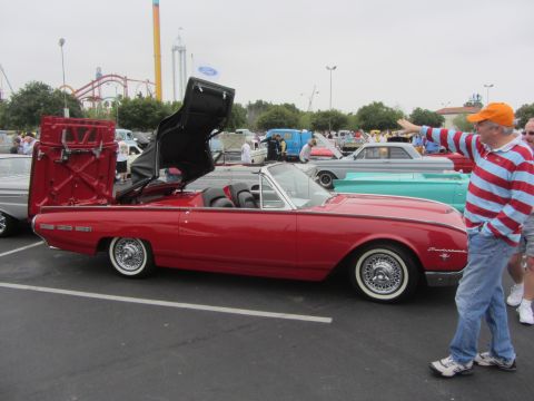 1964 Ford Ranchero - A red convertible's top is open, revealing its mechanism, as a man gestures towards it with other vehicles and rides visible.