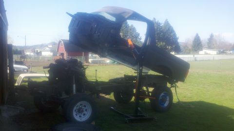 1988 Ford Bronco - Truck chassis and cab are disassembled and lifted by a hoist in a grassy yard.