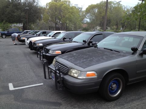 2004 Ford Crown Victoria - A line of Ford Crown Victoria police cars, including a gray one with blue wheel rims and a push bumper, are parked in a lot.
