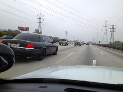 2004 Ford Crown Victoria - A black Ford Crown Victoria is seen driving on a highway with other vehicles under a cloudy sky with power lines overhead.