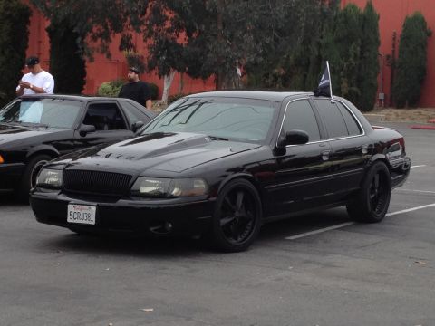 2004 Ford Crown Victoria - This black sedan, identified as a 2004 Ford Crown Victoria, features a modified hood and blacked-out wheels.