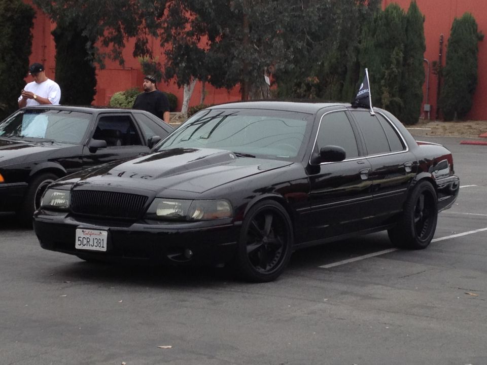 Black sedan with a custom hood and black rims, parked next to another car. A flag is on the roof