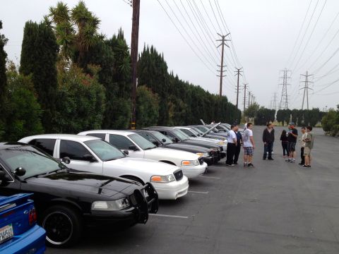 2004 Ford Crown Victoria - A line of police interceptor style sedans are parked in a lot, with several people standing around.