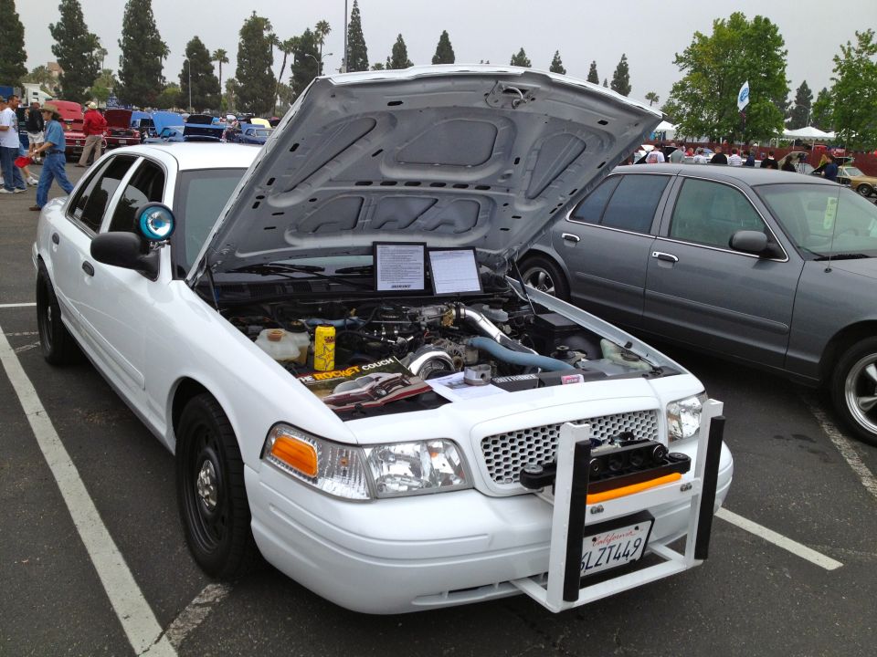 White police car with hood open, showing engine and a push bar. Another gray car is parked next to it