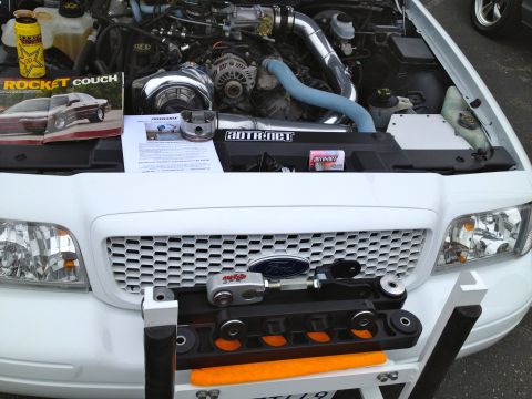 2004 Ford Crown Victoria - The engine bay of a white 2004 Ford Crown Victoria is open, revealing a modified engine with a turbocharger and custom piping.