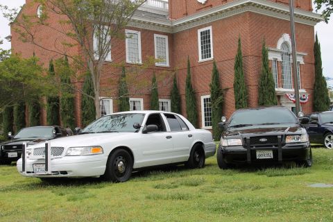 2004 Ford Crown Victoria - Two police cruisers, a white and a black model, are parked on a lawn in front of a large brick building.