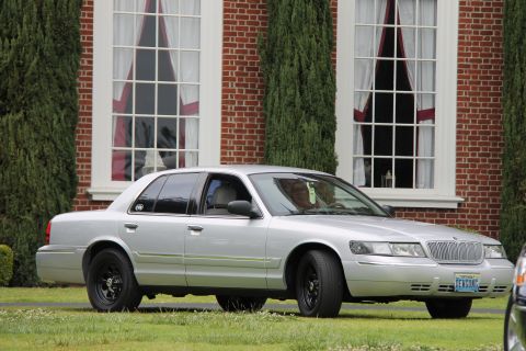 2004 Ford Crown Victoria - A silver 2004 Ford Crown Victoria is parked on a lawn in front of a brick building with large windows.