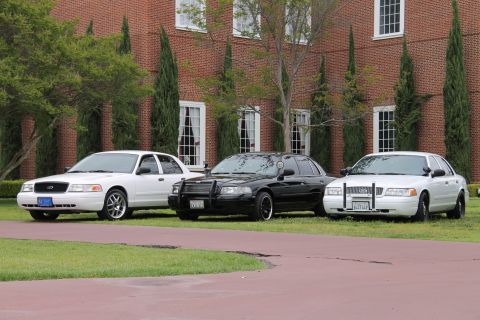 2004 Ford Crown Victoria - Three Ford Crown Victorias are parked in a row in front of a large brick building with tall windows.